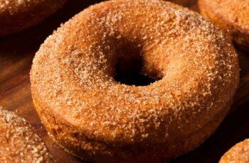 In the center of the image, a tantalizing display of Apple Cider Donuts steals the spotlight. These delectable treats are a warm, golden-brown hue, their surfaces adorned with a dusting of cinnamon-sugar that glistens in the soft, diffused natural light. The donuts are arranged neatly in a circular pattern, creating an inviting focal point.