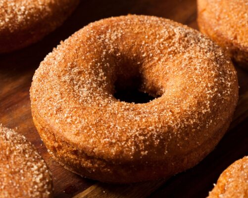 In the center of the image, a tantalizing display of Apple Cider Donuts steals the spotlight. These delectable treats are a warm, golden-brown hue, their surfaces adorned with a dusting of cinnamon-sugar that glistens in the soft, diffused natural light. The donuts are arranged neatly in a circular pattern, creating an inviting focal point.
