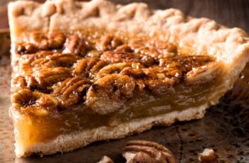 a freshly baked pecan pie takes center stage. The pie rests in a golden-brown, flaky pastry crust, its edges crimped to perfection. The surface of the pie is a rich, glossy caramel hue, a testament to the sugary magic that has occurred during its time in the oven.
