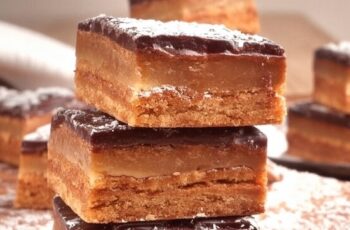 This image showcases delectable Millionaire's Shortbread bars, stacked enticingly on a wooden board. Each piece features three distinct layers: a buttery shortbread base, a smooth and chewy caramel center, and a glossy, firm chocolate topping. Lightly dusted with powdered sugar, these treats are both visually appealing and mouthwatering, making them a perfect dessert for any occasion. Ideal for sharing at parties or enjoying as a sweet indulgence, these bars highlight the perfect balance of flavors and textures that Millionaire's Shortbread is known for.